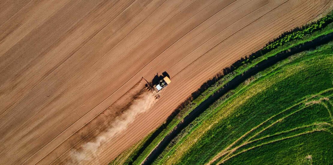 Aerial view of a tractor ploughing a brown field next to a green pasture.